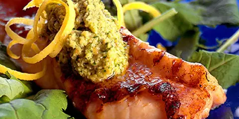 Grillad havsabborre med en grön topping och citronskal, serverad på en bädd av gröna blad.