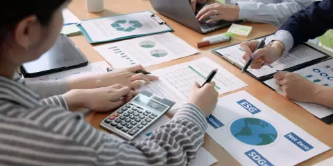Des mains travaillent sur des documents financiers, une calculatrice et un ordinateur portable. L'ergonomie organisationnelle est au cœur de cette réunion axée sur le développement durable.