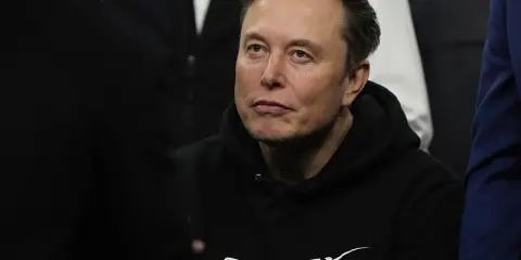 Elon Musk w bluzie SpaceX, zamyślony, w otoczeniu innych osób.