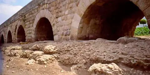 Antiguo puente de piedra con arcos, que es un puente de gran tamaño y robustez, con ruinas a sus pies.