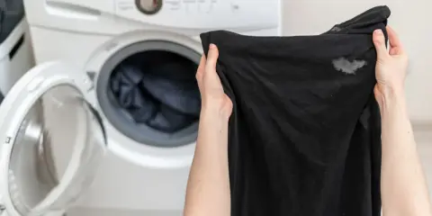 Manos sostienen ropa negra con manchas blancas, cerca de una lavadora abierta. ¿Por qué la ropa negra sale con manchas blancas?
