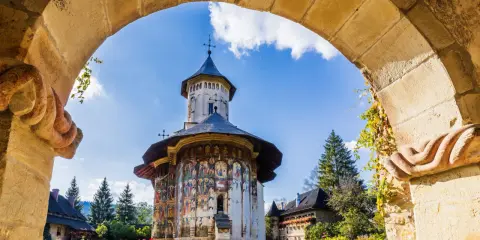 Malowany klasztor w Bukowinie, Rumunia, widziany przez kamienny łuk. Kolorowe freski zdobią ściany cerkwi, a wokół kwitną róże.