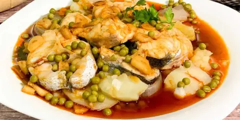 Merluza a la gallega con patatas, guisantes y ajo frito en una salsa rojiza.