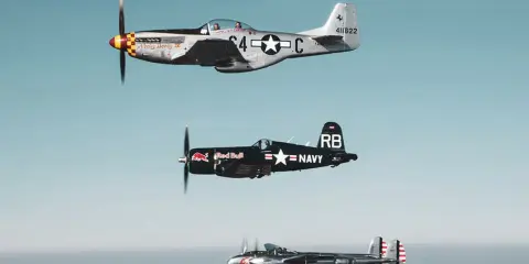 Drei historische Flugzeuge im Flug: eine silberne P-51 Mustang, ein schwarzer Red Bull Flieger (Corsair) und eine P-38 Lightning.