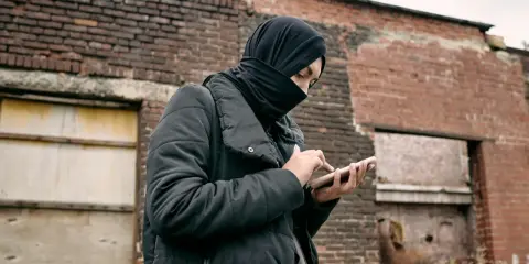 Osoba w czarnej chuście i kurtce trzyma telefon na tle zrujnowanej ceglanej ściany.