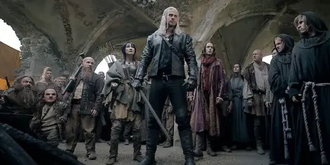 Geralt z Rivii stoi pośród tłumu, gdy ścieżki przeznaczenia w Wiedźminie splatają się. Widać krasnoludy, ludzi i zakapturzonych mnichów.