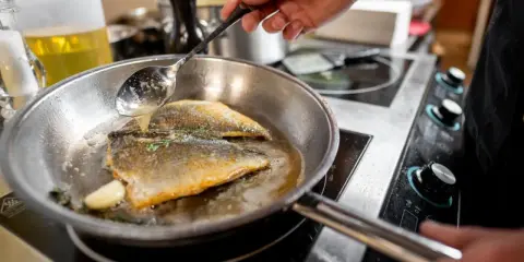 Un chef cocina pescado en una sartén de acero inoxidable, bañándolo con aceite caliente.