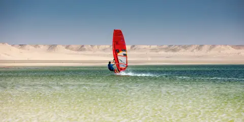 Windsurfer mit rotem Segel gleitet über das Wasser bei Dakhla, Marokko, vor einer Wüstenlandschaft.
