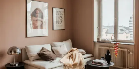 Un divano bianco accogliente con cuscini e una coperta color **rosa pastello** su un tappeto soffice. Alle pareti, due quadri e una finestra con vista.