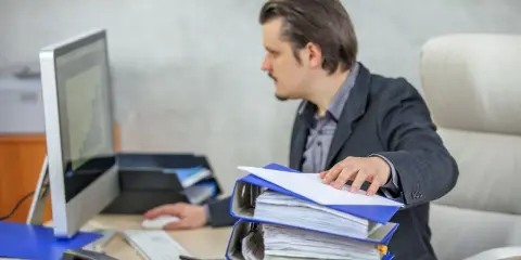 Mężczyzna w garniturze pracuje przy biurku, przeglądając dokumenty. Przed nim stos segregatorów, komputer i czerwony telefon. To obraz codziennego postępowania administracyjnego.