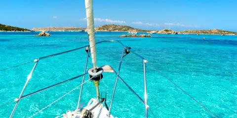 La prua di una barca a vela naviga verso La Maddalena, tra acque cristalline e isole rocciose.