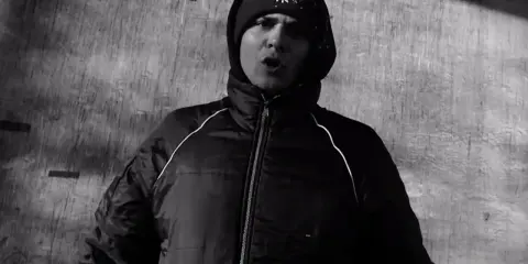 Młody mężczyzna w czapce z daszkiem i kurtce, z ustami otwartymi w geście mówienia, przypomina belmondawg hau.