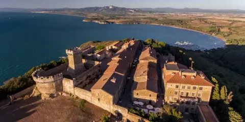 Borgo medievale fortificato domina la campagna toscana lungo il mare, con un castello imponente e case dai tetti rossi affacciate su un golfo con barche a vela.