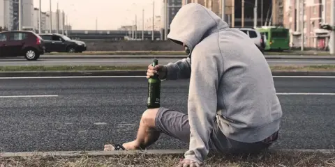 Mężczyzna w kapturze siedzi przy drodze z butelką piwa. Jego postawa sugeruje walkę z nałogiem, być może potrzebuje pomocy terapeuty od uzależnień.