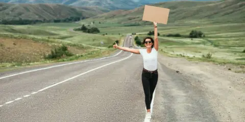 Kobieta z kartonem i kciukiem w górze na pustej drodze. Uczy się, jak łapać stopa, podróżując przez malownicze krajobrazy.