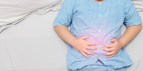 Niño con pijama azul, acostado en cama blanca, se agarra la barriga con las manos. Se visualiza un área rosada en su abdomen, indicando dolor barriga niños.