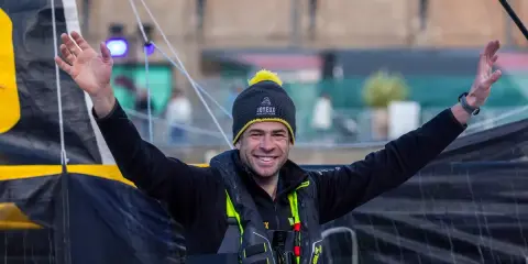 Nicolas d'Estais, marin souriant, bras levés, porte un bonnet et un gilet de sauvetage, prêt pour l'aventure en mer.