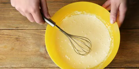 Mains préparant la pâte à crêpes dans un bol jaune. La pâte est prête pour son temps de repos.
