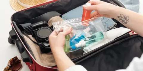 Ręce pakują kosmetyki w przezroczystej kosmetyczce do czerwonej walizki, obok słomkowego kapelusza i okularów.