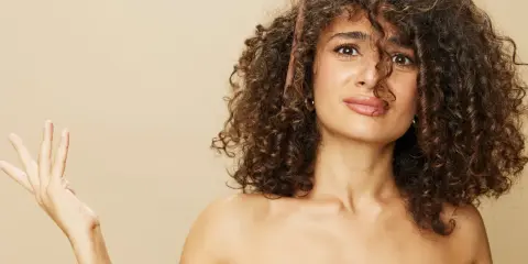 Une femme aux cheveux longs et bouclés, une brosse à cheveux coincée dedans, lève une main ouverte.