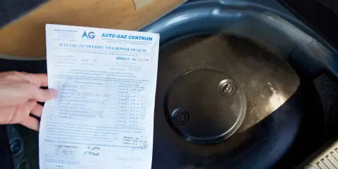 Fragment dokumentu z danymi homologacji butli LPG obok okrągłej pokrywy w bagażniku samochodu.
