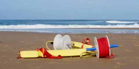 Deux bobines de fil, une blanche et une rouge, sont attachées à un tube de sauvetage jaune sur une plage.