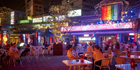 A vibrant night scene in gay Taipei, with people dining outdoors under colorful lights and a rainbow flag.