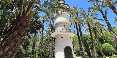 Torre blanca con balcón y puerta verde, rodeada de palmeras y vegetación exuberante. Un lugar encantador que ver en Elche.