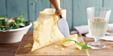 Ein Stück Parmesan, perfekt für vegetarische Gerichte, wird mit einem Käsemesser angeschnitten. Daneben ein Glas Weißwein und ein Salat.