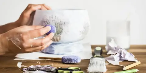 Ręce ozdabiające wazon techniką decoupage. Na stole nożyczki, pędzel, serwetki z lawendą i klej.