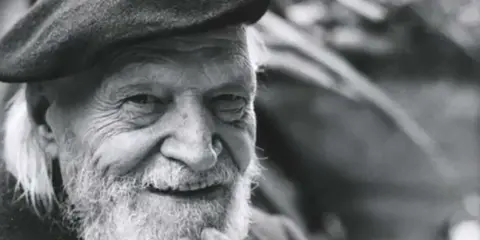Un anziano con barba e berretto, il suo sorriso saggio evoca il significato che la morte si sconta vivendo appieno ogni istante.