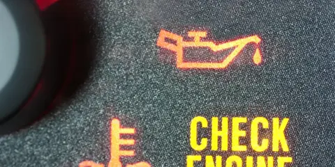 Czerwona kontrolka oleju świeci się na desce rozdzielczej, sygnalizując problem. Obok widoczny symbol termometru i napis "CHECK ENGINE".