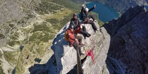 Trzech alpinistów w kaskach siedzi na szczycie skalistego Mnicha, machając do kamery. W tle rozciąga się malowniczy widok na Tatry i jezioro.
