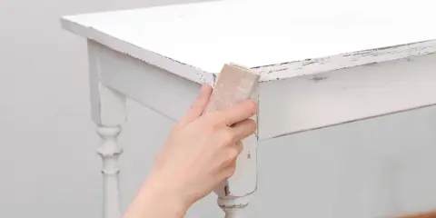 Main tenant, comment décaper un meuble peint. Une main utilise du papier de verre pour poncer le bord d'une table blanche écaillée.