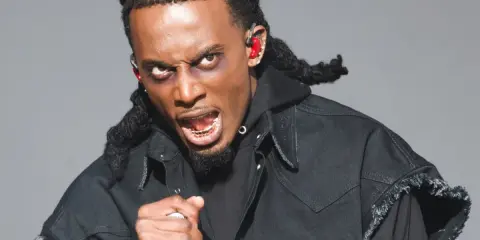 Playboi Carti z mikrofonem, w czarnej kurtce i z charakterystycznymi warkoczami. Jego ekspresja sugeruje energię znaną z jego playboi carti utworów.