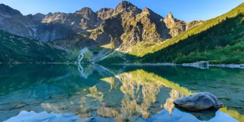 Gdzie w Tatry? Jezioro Morskie Oko odbija skaliste szczyty i zielone zbocza. Słońce oświetla góry, tworząc magiczny widok.
