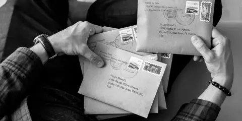 Ręce trzymające stos brązowych kopert, każda z adresem i znaczkiem pocztowym. Wygląda jak przygotowanie do wysyłki listów poleconych ekonomicznych.