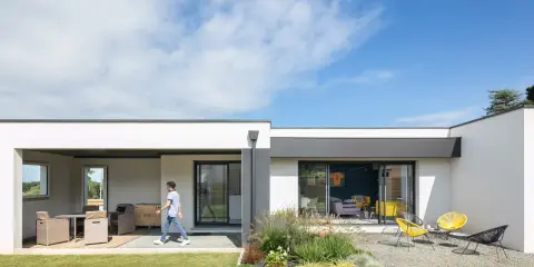 Une jeune personne traverse la pelouse devant une petite maison moderne avec une terrasse aménagée et un jardin de gravier.