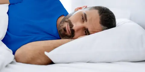 Hombre con barba en cama, con ojos abiertos, vistiendo camiseta azul. Duermo pero no descanso, ¿qué puedo hacer?