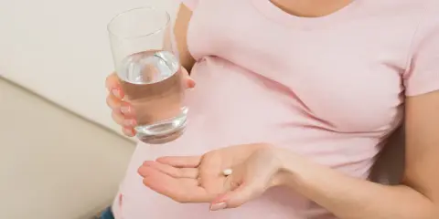 Kobieta w ciąży trzyma tabletkę i szklankę wody, szukając odpowiedzi na pytanie: co na ból głowy w ciąży.