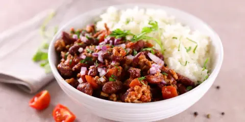 Un bol de riz blanc et de chili con carne recette de chef, garni d'oignons rouges et de persil. Des tomates cerises et du poivre en grains décorent la table.
