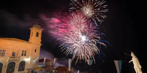 Spettacolo pirotecnico illumina il cielo notturno per la festa di San Marino. Folla ammira i fuochi d'artificio da una piazza con un edificio storico.