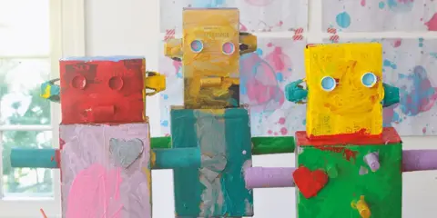 Drei bunte Roboter, die Kinder gebastelt haben, stehen nebeneinander. Sie sind aus Kartons und Rollen gefertigt und mit leuchtenden Farben bemalt.