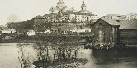 Widok na miasto z rzeką i kościołem. W oddali widać potężną budowlę sakralną z kopułami. Na pierwszym planie rzeka z wysepką i drzewami. Po prawej stronie drewniane budynki nad wodą. Ktoś pisze na Berdyczów.