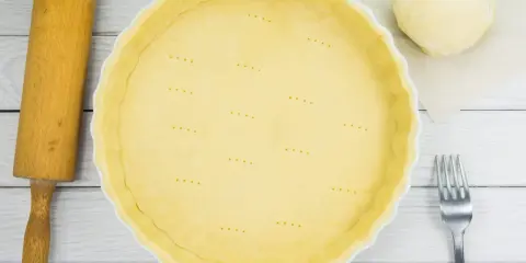 Une pâte brisée est prête dans une casserole blanche, avec une boule de pâte, une rouleau et une fourchette à côté.