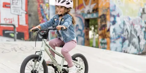 Uśmiechnięta dziewczynka w kasku radośnie pedałuje na swoim pierwszym rowerze dla dziecka. Tło to kolorowe murale.