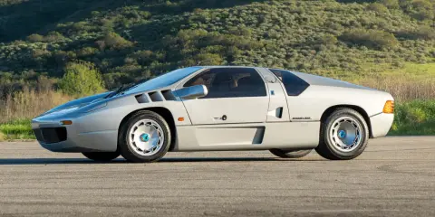 Silberner Isdera Imperator 108i mit markanten Lufteinlässen und futuristischem Design, geparkt vor einer grünen Hügellandschaft.