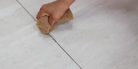 Main d'une personne nettoyant le joint de carrelage avec une éponge. La couleur du joint est sombre, contrastant avec le carrelage clair.