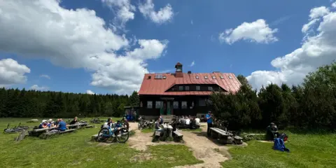 Chatka górzystów z czerwonym dachem, otoczona lasem. Ludzie odpoczywają przy stołach, obok stoją rowery.