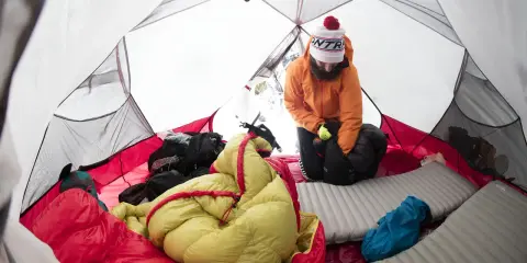 Wnętrze namiotu z kolorowymi śpiworami i sprzętem. Mężczyzna w pomarańczowej kurtce przygotowuje się do snu pod namiotami.